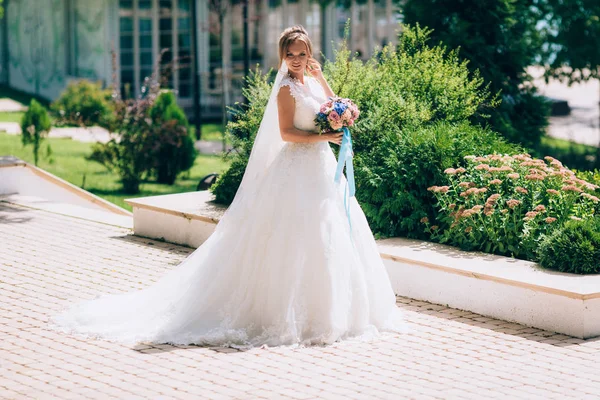 Die Braut trägt ein kurviges Kleid und geht mit einem langen Schleier die malerische Allee entlang. Hochzeitsspaziergang mit einem Blumenstrauß. — Stockfoto