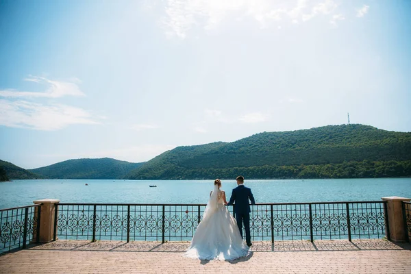 Nygifta stå på bryggan och beundra bukten och flytande båtarna. En flicka i en magnifik brudklänning och en man i en strikt kostym står vid vattnet och titta på de gröna kullarna. — Stockfoto