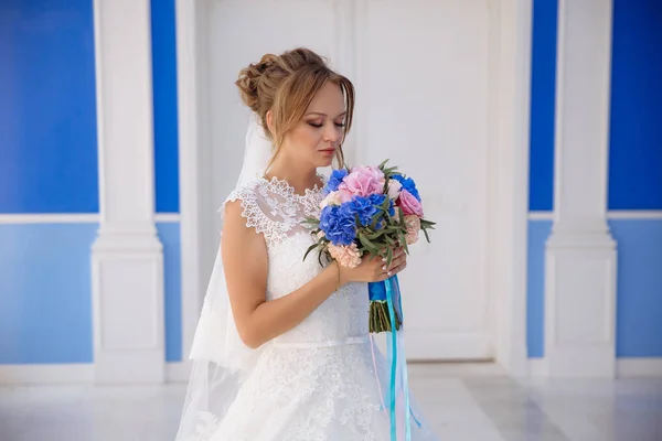 La mariée blonde se tient sur un bâtiment bleu et bleu dans une robe de mariée et profite de l'arôme d'un bouquet de fleurs. La fille renifle roses et pivoines . — Photo