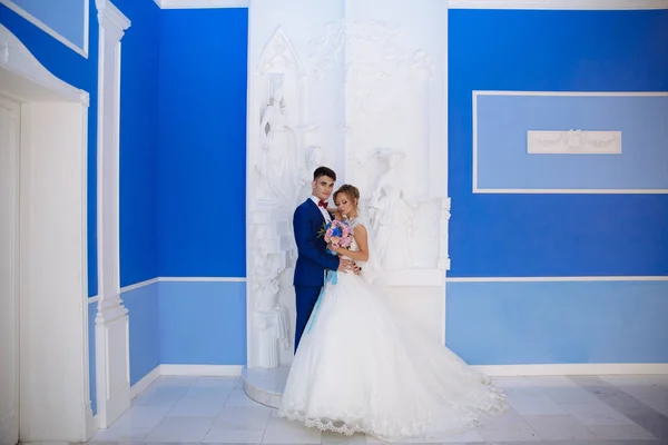 The bride and groom are posing for a photo in a huge blue hall with white columns. The color of the grooms costume is combined with the color of the walls and the bouquet of the bride.