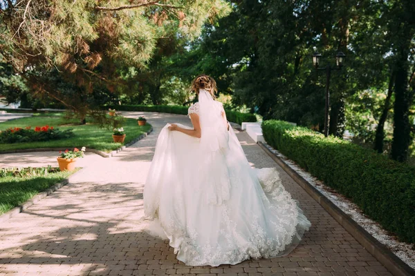 A loira em um vestido de noiva e com um véu corre ao longo da avenida verde. A menina se apressa para conhecer seu noivo . — Fotografia de Stock