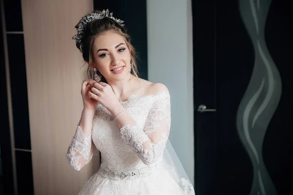 A noiva em um vestido branco rendado e véu está indo para a cerimônia. Retrato de uma menina colocando brincos e sorrindo . — Fotografia de Stock