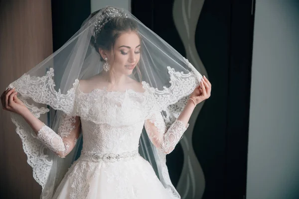 Porträtt av bruden i en spets bröllopsklänning och en lång slöja. Flickan ser på sin outfit, hennes hår är dekorerad med en krona, i hennes öron stora blanka örhängen. — Stockfoto