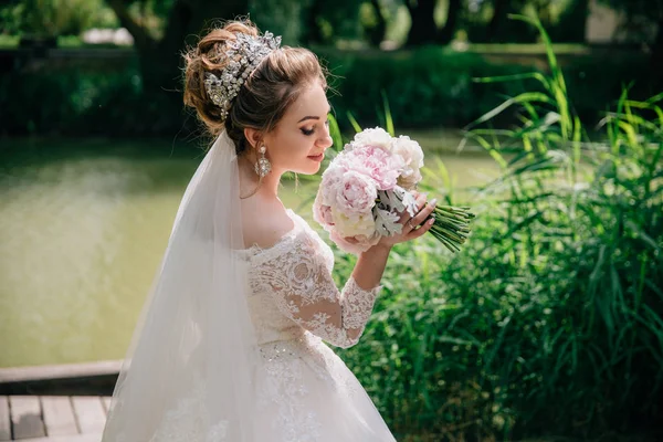 Vista desde atrás de la novia con el pelo recogido en un gran bulto con un velo olfatea su ramo de bodas de peonías y sonríe tiernamente . —  Fotos de Stock