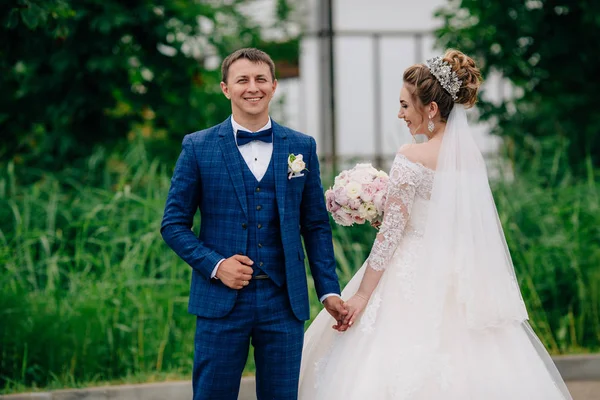 Um homem sorridente em um terno de negócios elegante sorri e mantém sua noiva, que fica no vestido de noiva com as costas . — Fotografia de Stock