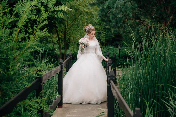 La novia en el vestido de novia está de pie al principio del puente sobre el río en un bosque verde. La chica admira el río abajo y quiere cruzarlo . —  Fotos de Stock