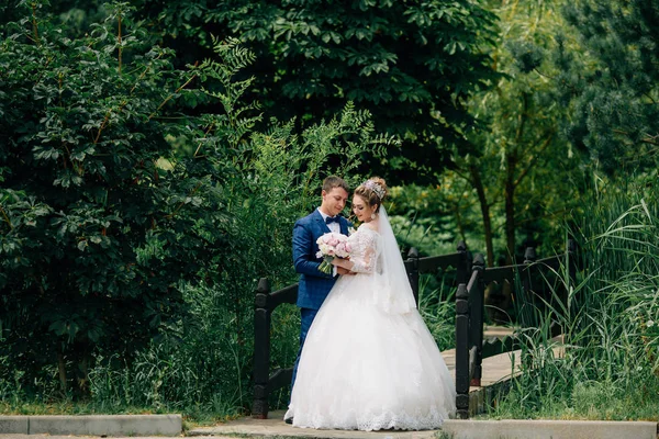 A noiva e o noivo acabaram de se casar e caminham no parque, posando perto da ponte do outro lado do rio. Os recém-casados abraçam e admiram o buquê de flores . — Fotografia de Stock