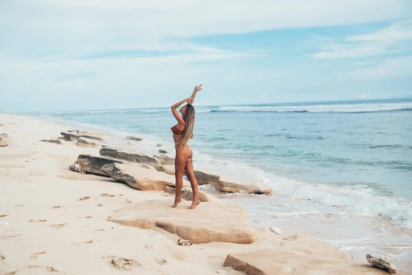 Зовні тонка витончена модель жінки на порожньому білому пляжі на тлі оливкового океану. Засмагла спортивна дівчина позує вбік, показує стрункі ноги і грубу дупу, тягне руки до — стокове фото