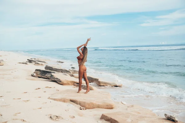 Lá fora, uma mulher magro gracioso modelo em uma praia branca vazia contra um fundo do oceano de azeitona. Menina atlética bronzeada posa de lado, mostra pernas finas e uma bunda áspera, desenha as mãos para o — Fotografia de Stock