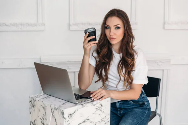 Eine schöne schlanke Kaffeeliebhaberin mit grau glänzenden Augen sitzt in einem gemütlichen Café hinter einem Laptop und wartet auf eine Nachricht von einem Freund. — Stockfoto