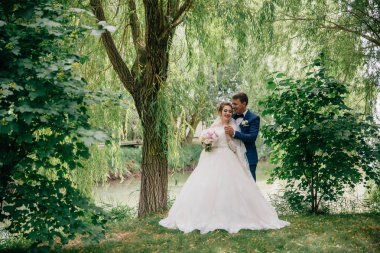 Gelin ve damat ağaçlar ve bir su birikintisi arka plan karşı poz olan. Bir adam onun sevgili arkadan sarılarak ve elini sıkıca tutar. Güzel düğün buketi kız elinde.