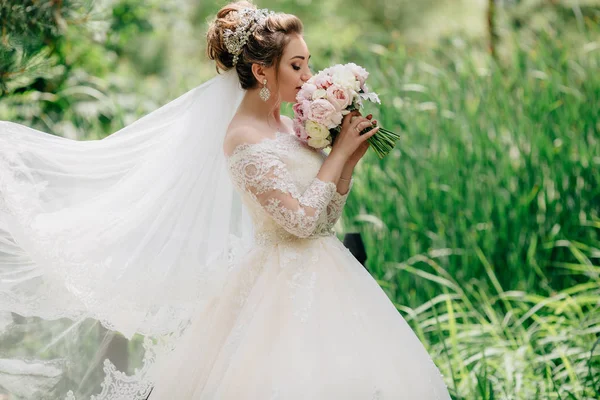 La novia se encuentra en un campo con hierba verde alto y disfruta del aroma de su ramo de flores de la boda. La moda de las chicas se desarrolla en las ráfagas de un viento de verano . —  Fotos de Stock