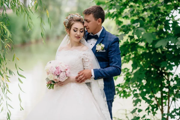 En man från bakom kramar sin älskade. Flickan täcker ögonen, hon känner sig glad och lugn på bröllopsdagen. Ung poserar i en grön park. — Stockfoto