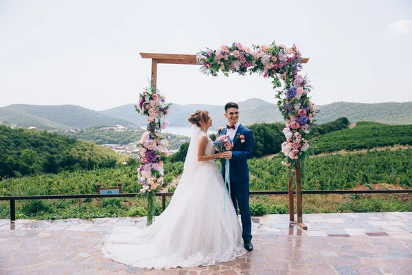 Marido e esposa estão de pé sob um arco de flores em meio a uma natureza incrivelmente bonita. O cara olha para seus amigos e sorri para eles, a menina olha para o marido apaixonado . — Fotografia de Stock