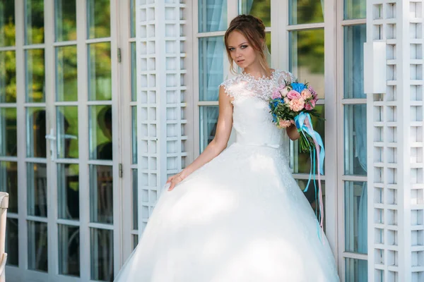 Novia en un vestido cerrado posando contra una vidriera. La niña sostiene un vestido con una mano, y el segundo ramo de flores con cintas azules . —  Fotos de Stock