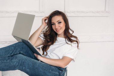 Basit şık kıyafetlerle çekici Avrupa görünümlü kız laptop ile oturup uzun kıvırcık saçlı portresi.