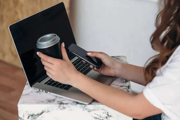 Nahaufnahme der Hand eines jungen Mädchens, das ein Glas Kaffee und ein Smartphone mit leerem Bildschirm in der Hand hält, vor sich ein Laptop auf dem Schreibtisch. moderne Technologie, Geräte, Menschen-Konzept. — Stockfoto