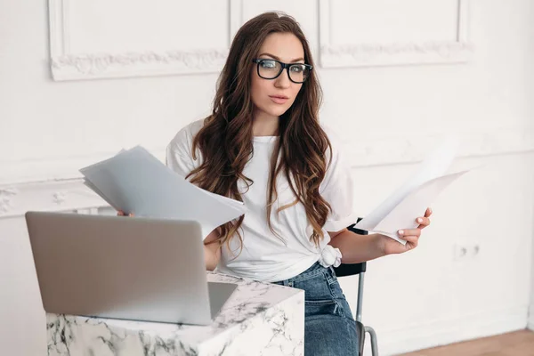 Eine junge schöne Frau reibt sich in einem modernen Büro, das mit Papierkram beschäftigt ist. Sie sitzt an einem Tisch mit einem Notizbuch und studiert den Text der Berichte. attraktive Europäerin mit welligem Haar in Brille. — Stockfoto