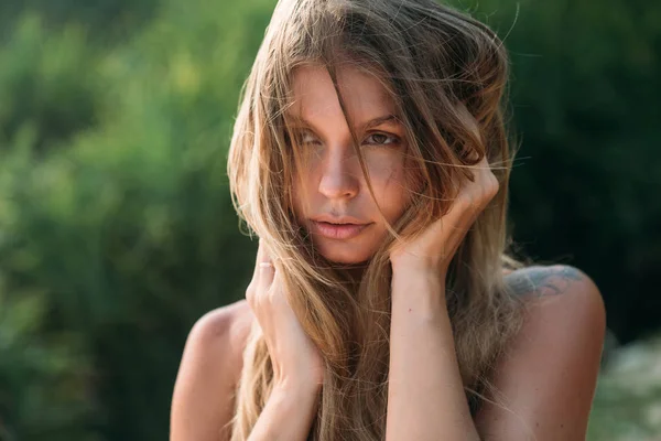 A close view of a beautiful tender sweet girl starts her hands in her hair, looks into the distance and dreams. A model with bare shoulders and brown eyes, the wind blows her hair. — Stock Photo, Image