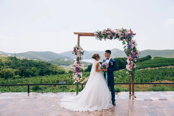 Jonggehuwden verliefd kijken elkaar en genieten van de trouwdag. Ze staan op een boog van roze, wit en blauwe bloemen. — Stockfoto