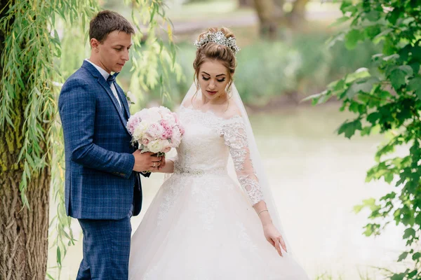 Um homem em um terno xadrez admira seu vestido de noiva de esposas e ajuda-a a manter um buquê de peônias. A noiva corrige a saia magnífica de seu vestido . — Fotografia de Stock