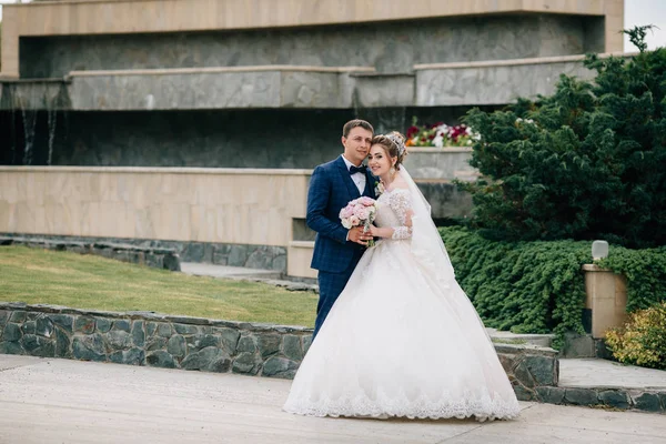 Bruden och brudgummen omfamna och promenera i parken. En man kramar sin älskade, och flickan glatt ser på kameran och visar sin brudbukett till sina vänner. — Stockfoto