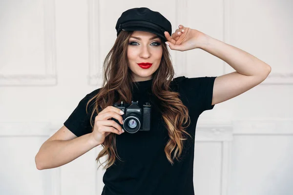 A beautiful young girl, has a clean healthy face, long silky hair, a bright make-up, is dressed in a black cap and t-shirt, holds a camera in her hand.