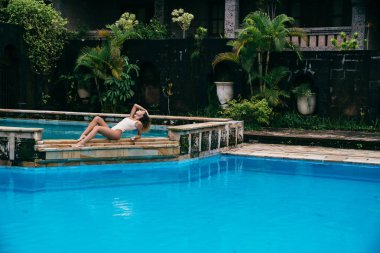 Bir bikini güneşlenme ve Yüzme Havuzu mavi su ile yakınındaki yalanlar güzel kız. Seksi model spor şekil ile istirahat ediyor. Hafta sonları bir mayo kıvırcık saçlı genç kadın.