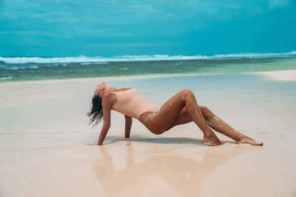 Slender girl with sexy ass posing on the beach in a swimsuit. A girl with a beautiful body enjoys the ocean and the sun. A model with short dark curly hair posing on white sand. — Stock Photo, Image