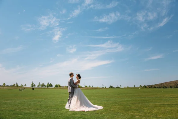 Sposi stanno camminando lungo il campo verde del golf club in un giorno di nozze. Lo sposo in un abito da lavoro è grigio e la sposa in un abito bianco di lusso con un velo si tiene per mano . — Foto Stock