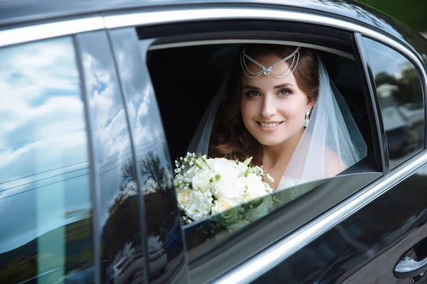 Uma vista do lado de fora do carro, uma jovem noiva europeia senta-se no banco de trás de uma limusine preta, segura um buquê de casamento na mão e sorri . — Fotografia de Stock