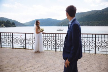 Genç bir adam onun gelin gelinlik bir nehir ve bir tekne yelken hangi tepeler yeşil ormanıyla kaplı zemin karşı bakar. Düğün günü doğada güzel bir yerde.