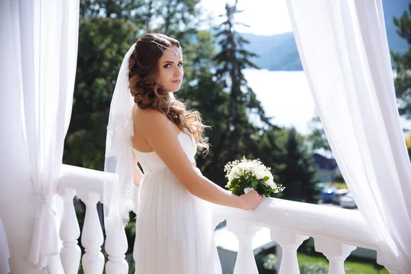 Темноволоса, кучерява дівчина стоїть на балконі з видом на річку, перетворюється в камеру, одягнену в красиву весільну сукню, завісу . — стокове фото