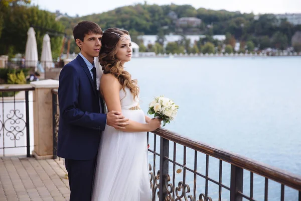 Una novia embarazada con un vestido de novia está de pie en el muelle, admirando la vista del río, detrás de sus abrazos a su marido. Las relaciones armoniosas, la boda, los amantes del concepto . — Foto de Stock