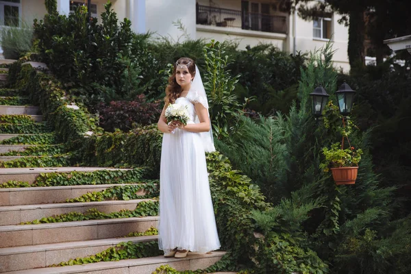 A bela noiva jovem fica em escadas de restolho em um vestido de estilo grego, seu cabelo é encaracolado, decorado com contas, ela está segurando um buquê de casamento de rosas brancas. . — Fotografia de Stock