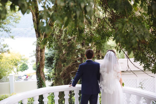 Une femme avec un voile de dentelle blanche et une robe de mariée tient un homme en costume d'affaires, un couple se tient sur le balcon et admire la vue sur le jardin et la rivière . — Photo