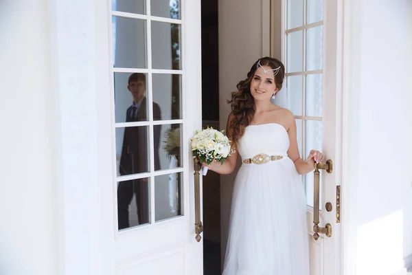 A noiva fica à porta do salão de casamento, sorri e chama seu homem para ir com ela. A silhueta do cara é refletida nas portas . — Fotografia de Stock