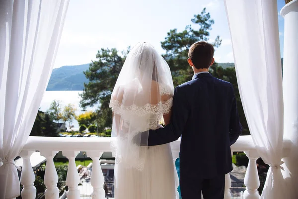 Die Aussicht von hinten, das Brautpaar steht auf dem Balkon ihres schneeweißen Schlafzimmers und bewundert den Blick auf den Fluss, den Garten und den blauen Himmel. die Braut in Brautkleid und Schleier — Stockfoto