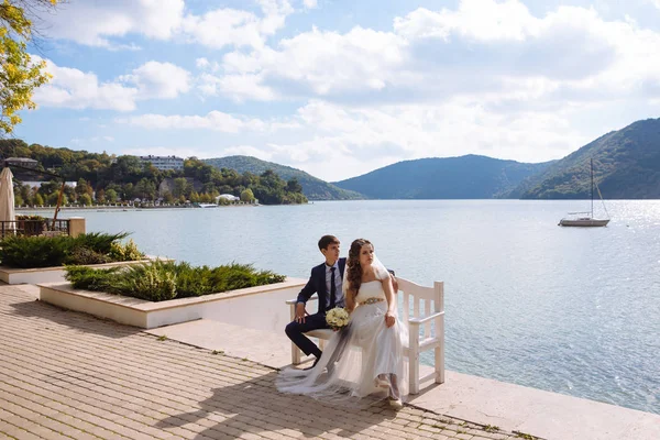 Un hombre de negocios con estilo y una chica en un vestido de estilo griego sentarse en el paseo marítimo, admirar la vista del río, son felices en el día de su boda para ver tanta belleza alrededor . —  Fotos de Stock