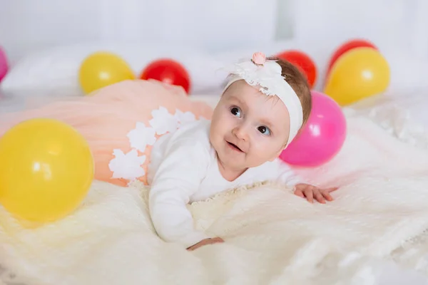 The baby is lying on the bed and looking away with interest and a smile. The girl is dressed in a beautiful dress and crawls on a white bed. — Stock Photo, Image