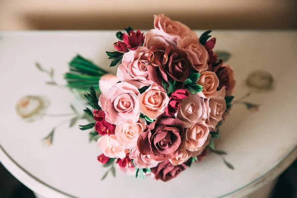 A wedding bouquet of red and pink roses lies on a wooden vintage table. Golden rings of newlyweds on flowers. The concept of wedding floristry and jewelry. — Stock Photo, Image