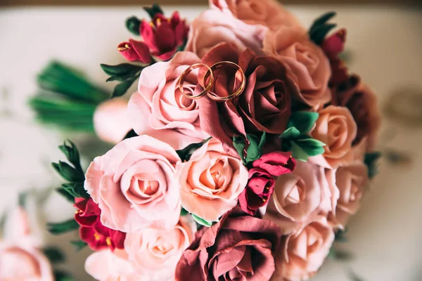 Un ramo de boda de rosas rojas y rosas se encuentra en una mesa vintage de madera. Anillos dorados de cerca de recién casados en flores. El concepto de boda floristería y joyería . — Foto de Stock