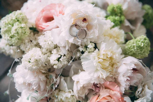Anillos dorados de cerca de recién casados en flores. Un ramo de bodas de hermosas y frescas peonías se encuentra en una mesa vintage de madera. El concepto de boda floristería y joyería . — Foto de Stock