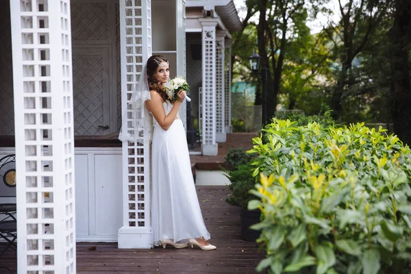 Una chica con un vestido blanco claro y un velo sostiene un ramo de bodas y posa cerca del porche blanco de una casa de un solo piso . — Foto de Stock