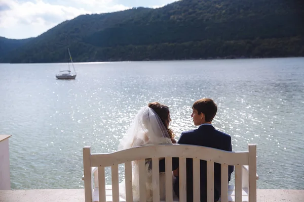 Un uomo in un abito elegante costoso è seduto su una panchina con la sua fidanzata in un velo e un vestito bianco, ammirando l'ampio fiume lungo il quale le navi e gli yacht nuotano. . — Foto Stock