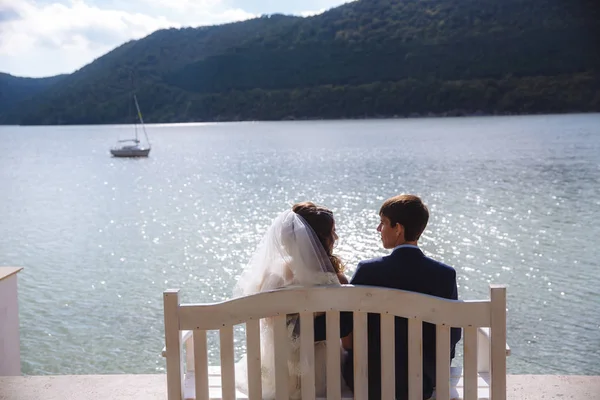 Un hombre en un traje caro elegante está sentado en un banco con su prometido en un velo y un vestido blanco, admirando el ancho río a lo largo del cual los barcos y yates nadan . —  Fotos de Stock