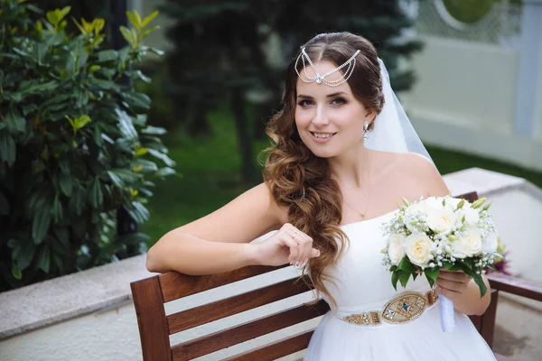 Усміхнена наречена одягнена в весільну сукню в грецькому стилі, її волосся вкриті лалатином, тримає в руці букет білих квітів, посміхається, сидячи в парку . — стокове фото