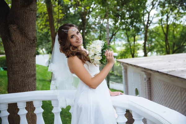 Jeune belle mariée avec de longs cheveux noirs bouclés posant dans une robe de mariée sur un balcon blanc, souriant, tenant un bouquet de fleurs dans sa main . — Photo