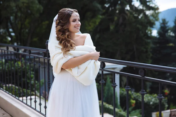 Une belle jeune mariée aux longs cheveux foncés, flânant le long du remblai, revêtue d'un manteau de fourrure pour ne pas geler, sourit joyeusement . — Photo