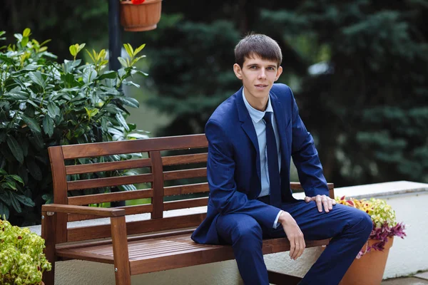 A young lean guy, the student is sitting on a bench in the park, today he is a jeweler and very happy. The groom wears a classic shadow suit and a blue tie.